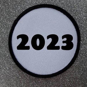 Könnte beinhalten: Ein kreisförmiger Aufnäher mit hellblauem Zentrum und schwarzem Rand. Die Zahl "2023" ist in fetter schwarzer Schrift über die Mitte des Aufnähers abgebildet. Der Hintergrund hat eine silberne Glitzertextur.