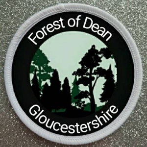 Könnte beinhalten: Runder weißer Aufnäher mit schwarzem Rand und weißem Text "Forest of Dean Gloucestershire" mit einer Silhouette von Bäumen in der Mitte.