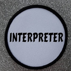 Può includere: Toppa rotonda bianca con cuciture nere attorno al bordo. Il testo "INTERPRETER" è stampato in nero sulla toppa.