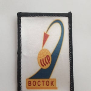 Op de afbeelding: Een witte geborduurde patch met een zwarte rand. De patch toont een blauwe en gele raket met een rode pijl en de tekst "BOSTOK" in geel.