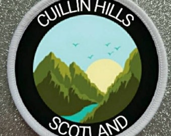 Peut inclure: Écusson rond blanc avec une bordure noire et le texte "Cuillin Hills Scotland". Le centre présente un paysage de montagne vert avec une rivière bleue et un soleil jaune.