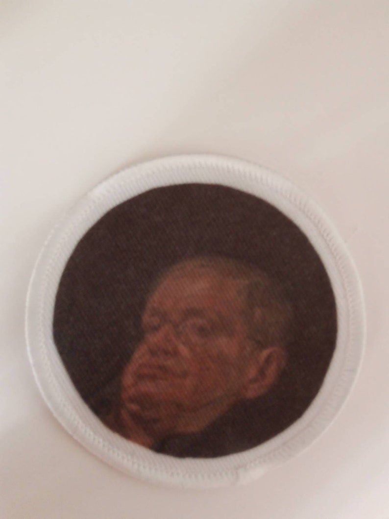 Op de afbeelding: Een witte stoffen patch met een bruine rand en een portret van een man met een bedachtzame uitdrukking. De man draagt een donker pak en heeft een lichte huidskleur.