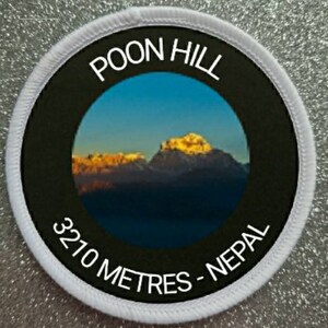 Może przedstawiać: Biała okrągła naszywka z czarnym tekstem, który brzmi "POON HILL 3210 METRES - NEPAL". Naszywka ma okrągły obraz pasma górskiego z niebieskim niebem i chmurami w tle.