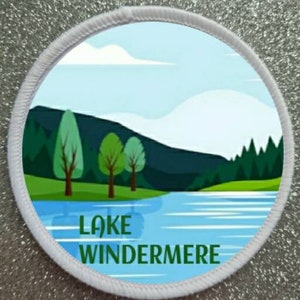 May include: Circular patch with a landscape design and the words "LAKE WINDERMERE". The image shows a lake, trees, and mountains under a blue sky. The patch has a white, stitched border.