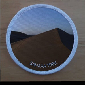 Może przedstawiać: Okrągła biała łatka z fotografią wydmy w Saharze. Tekst "SAHARA TREK" jest nadrukowany na łatce na biało.