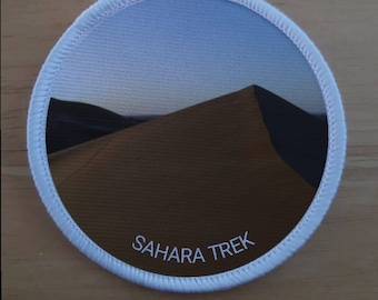 Może przedstawiać: Okrągła biała łatka z fotografią wydmy w Saharze. Tekst "SAHARA TREK" jest nadrukowany na łatce na biało.