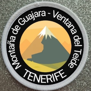 Puede incluir: Un parche circular con un gráfico de montaña. El parche presenta un borde negro con texto blanco que dice "Montaña de Guajara - Ventana del Teide TENERIFE". La montaña está representada en tonos de gris, blanco y naranja sobre un fondo amarillo.