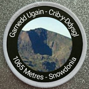 May include: A white and black circular patch with the text "Garnedd Ugain - Crib-y-Dysgl 1065 Metres - Snowdonia" around a photo of a mountain.