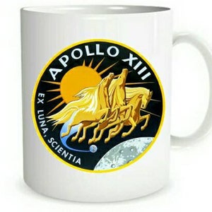 Op de afbeelding: Witte keramische mok met een zwart en geel Apollo XII-missie patch. De patch toont een gestileerde afbeelding van drie rennende paarden met een zon en maan op de achtergrond. De tekst "Apollo XII" staat in een zwarte cirkel met de tekst "Ex Luna, Scientia" in een zwarte boog onder de cirkel.