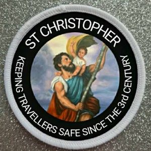 Könnte beinhalten: Runder weißer Aufnäher mit schwarzem Text und einem Bild des Heiligen Christophorus, der ein Kind trägt. Der Text lautet "St Christopher Keeping Travellers Safe Since The 3rd Century".