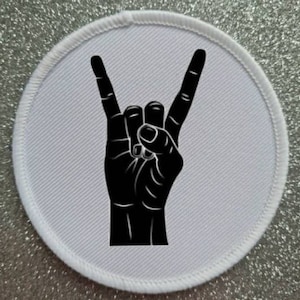 Może przedstawiać: Biała okrągła łatka z czarną sylwetką dłoni wykonującej gest rock and roll.