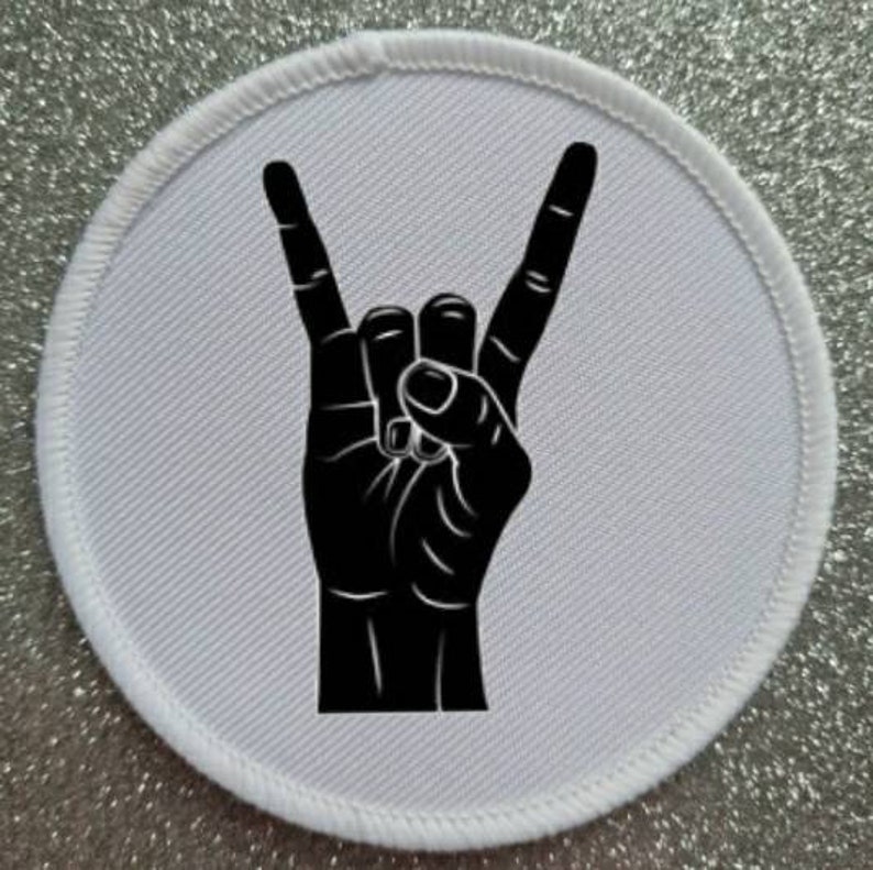 Może przedstawiać: Biała okrągła łatka z czarną sylwetką dłoni wykonującej gest rock and roll.