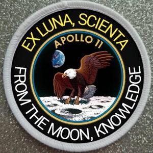 Pode incluir: Um remendo circular branco com um círculo preto no centro. O círculo preto tem uma águia branca com as asas abertas, em pé sobre uma superfície cinza com crateras. O texto "EX LUNA, SCIENTA" está em amarelo ao redor da parte superior do círculo preto. O texto "APOLLO 11" está em branco dentro do círculo preto. O texto "FROM THE MOON, KNOWLEDGE" está em branco ao redor da parte inferior do círculo preto.