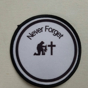 May include: A white round patch with a black border and the words "Never Forget" in a circle around a black silhouette of a person kneeling in front of a cross.