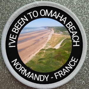 May include: Circular patch with the text "I'VE BEEN TO OMAHA BEACH NORMANDY - FRANCE" in white lettering on a black background. The center features a scenic view of a beach and coastline. The patch has a white border.