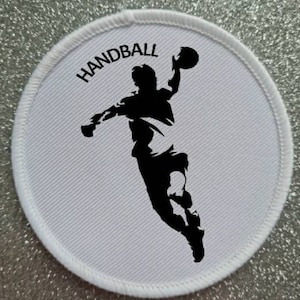 May include: White round patch with black stitching around the edge. The patch features a black silhouette of a handball player throwing the ball with the word "HANDBALL" printed in black above the player.