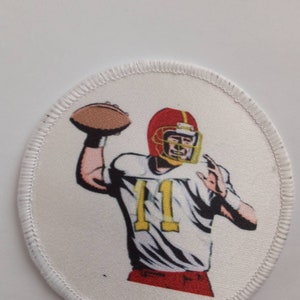 Könnte beinhalten: Runder Aufnäher mit einem Footballspieler in einem roten Helm und einem weißen Trikot mit der Nummer 11. Der Spieler wirft einen braunen Football. Der Aufnäher hat einen weißen Hintergrund und einen genähten Rand.