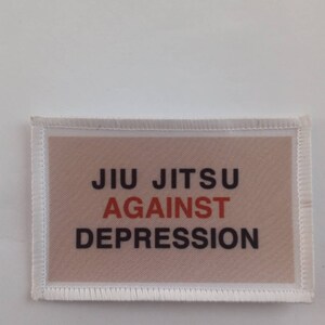 Op de afbeelding: Witte patch met een bruine achtergrond en de tekst "JIU JITSU AGAINST DEPRESSION" in zwarte en oranje letters.