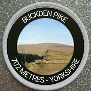May include: A white and black round patch with the text "Buckden Pike 702 Metres - Yorkshire" in white lettering. The patch features a photo of a landscape with a hill in the background.