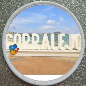 Può includere: Lettere bianche che compongono la parola "CORRALEJO" su una spiaggia sabbiosa con un oceano e un cielo blu sullo sfondo. Un piccolo fiore colorato si trova in primo piano.