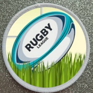 Puede incluir: Una pelota de rugby blanca y azul con el texto "RUGBY LEAGUE" en ella, sentada en hierba verde. La pelota está parcialmente oscurecida por un círculo blanco con fondo amarillo.