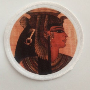 May include: A round fabric patch with a white border and a printed image of an Egyptian queen wearing a headdress with a cobra. The queen has dark hair, black eyes, and red lips. The background is a light orange color.