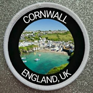 May include: White round patch with black border and text "CORNWALL ENGLAND, UK". The patch features a photo of a coastal town with a harbor and boats.