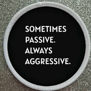Peut inclure: Un patch rond noir et blanc avec du texte blanc qui dit "Sometimes Passive. Always Aggressive."