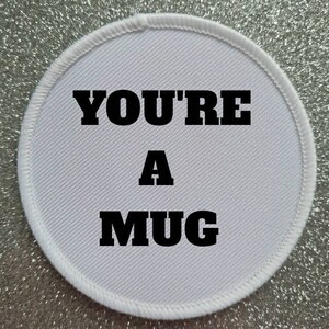 Op de afbeelding: Ronde witte patch met zwarte tekst die "You're a Mug" zegt.