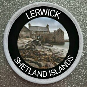 Puede incluir: Un parche redondo blanco con texto negro y un círculo negro en el centro. El texto dice "LERWICK SHETLAND ISLANDS". El círculo negro contiene una foto de un edificio de piedra con un cielo nublado en el fondo.