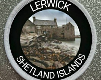 May include: A white round patch with black text and a black circle in the center. The text reads "LERWICK SHETLAND ISLANDS". The black circle contains a photo of a stone building with a cloudy sky in the background.
