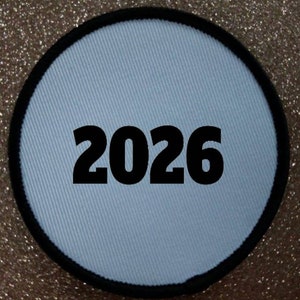 May include: A circular patch featuring a light blue fabric centre and a black border. The number "2026" is printed in bold black font across the centre. The background has a glittery gold effect.