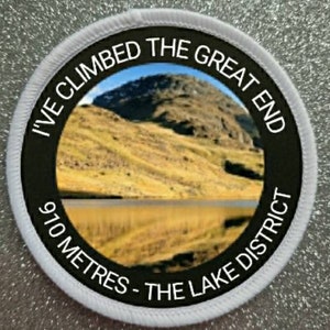 May include: White round patch with black text and a photo of a mountain and lake. The text reads "I've Climbed The Great End 970 Metres - The Lake District".