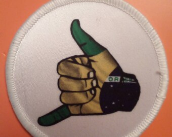May include: A white circular patch with a white border and a green, yellow, and black hand making the shaka sign. The hand is wearing a black wristband with white stars and the letters "OR" in white.