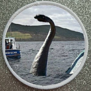 Pode incluir: Uma criatura preta e cinza semelhante ao monstro de Loch Ness com um pescoço longo emerge da água. Um pequeno barco com a palavra "DeepScan" no lado está no fundo.