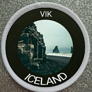 Pode incluir: Um remendo redondo branco com um círculo preto no centro. O círculo preto tem um círculo branco no centro com uma foto de uma praia de areia preta na Islândia. O texto "VIK" está no topo do círculo preto e "ICELAND" está na parte inferior.