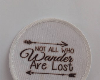 Może przedstawiać: Okrągła naszywka z białego materiału z brązowym haftem, na którym widnieje napis "Not all who wander are lost" z dwoma strzałkami po obu stronach tekstu.
