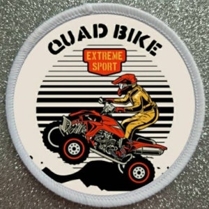 May include: Circular patch with a white border. It depicts a red quad bike and a rider in yellow protective gear. The words "QUAD BIKE" are above the image, with "EXTREME SPORT" in a red rectangle. Black and white stripes form a backdrop.