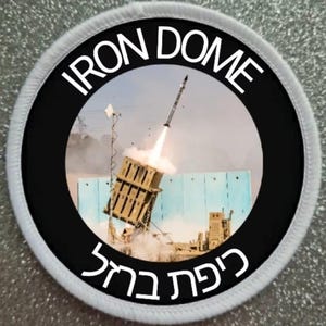 Puede incluir: Un parche circular blanco y negro con las palabras "IRON DOME" en letras blancas alrededor de un círculo negro. Dentro del círculo negro hay una foto de un lanzamiento de misiles con las palabras "כיפת ברזל" en hebreo.