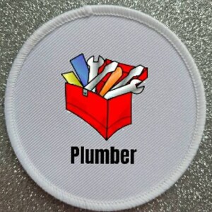 May include: A circular patch with a white border and a grey background. It displays a red toolbox graphic filled with tools. The word "Plumber" is printed in black beneath the toolbox.