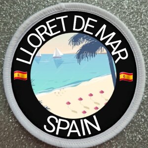 Può includere: Un cerotto rotondo bianco con un cerchio nero al centro. Il cerchio nero contiene il testo "LLORET DE MAR SPAGNA" in lettere bianche. All'interno del cerchio nero c'è una scena di spiaggia con palme, barche a vela e ombrelloni rosa su una spiaggia sabbiosa.