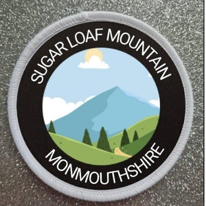 May include: White and black round patch with the text "Sugar Loaf Mountain Monmouthshire" and a cartoon illustration of a mountain with a blue sky and white clouds.