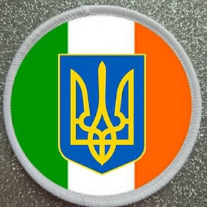 Puede incluir: Un parche redondo con el escudo de armas de Ucrania, un tridente dorado sobre fondo azul, centrado en un fondo blanco con rayas verdes y naranjas a cada lado.
