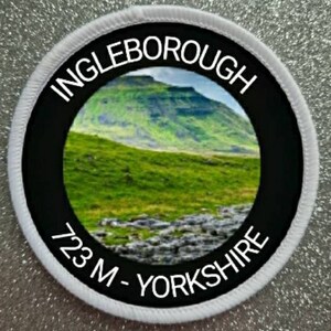 May include: White round patch with black border and white text. The text reads "Ingleborough 723 M - Yorkshire". The patch features a photo of a green hill with a rocky foreground.
