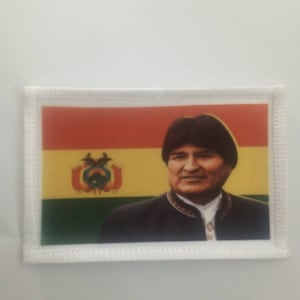 Peut inclure: Un écusson brodé blanc avec un drapeau bolivien rouge, jaune et vert et un portrait d'un homme portant un chapeau noir et une chemise blanche.