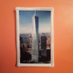 May include: A white framed patch featuring a photograph of the Freedom Tower in New York City. The tower is a tall, slender building with a pointed top. The photo is taken from a distance, showing the tower against a blue sky.