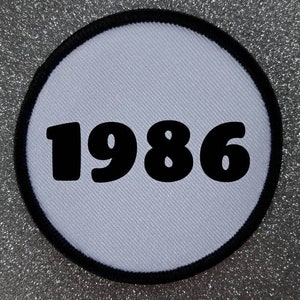 Op de afbeelding: Ronde witte patch met een zwarte rand en het jaar 1986 gedrukt in grote zwarte letters.