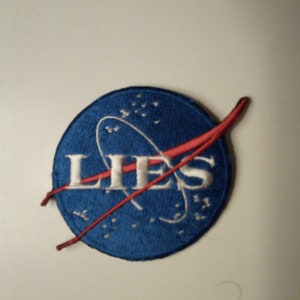 May include: A blue and red embroidered patch with the word "LIES" in white lettering. The patch is a parody of the NASA logo.