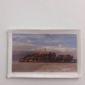 May include: A white fabric patch with a stitched border featuring a color photograph of Alcatraz Island in San Francisco Bay. The island is a former prison and is now a popular tourist destination.