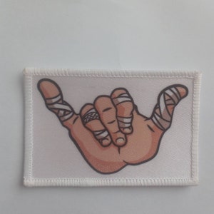 Puede incluir: Parche de tela blanca con una mano de dibujos animados haciendo el signo shaka. La mano está vendada con vendas blancas y tiene piel marrón.